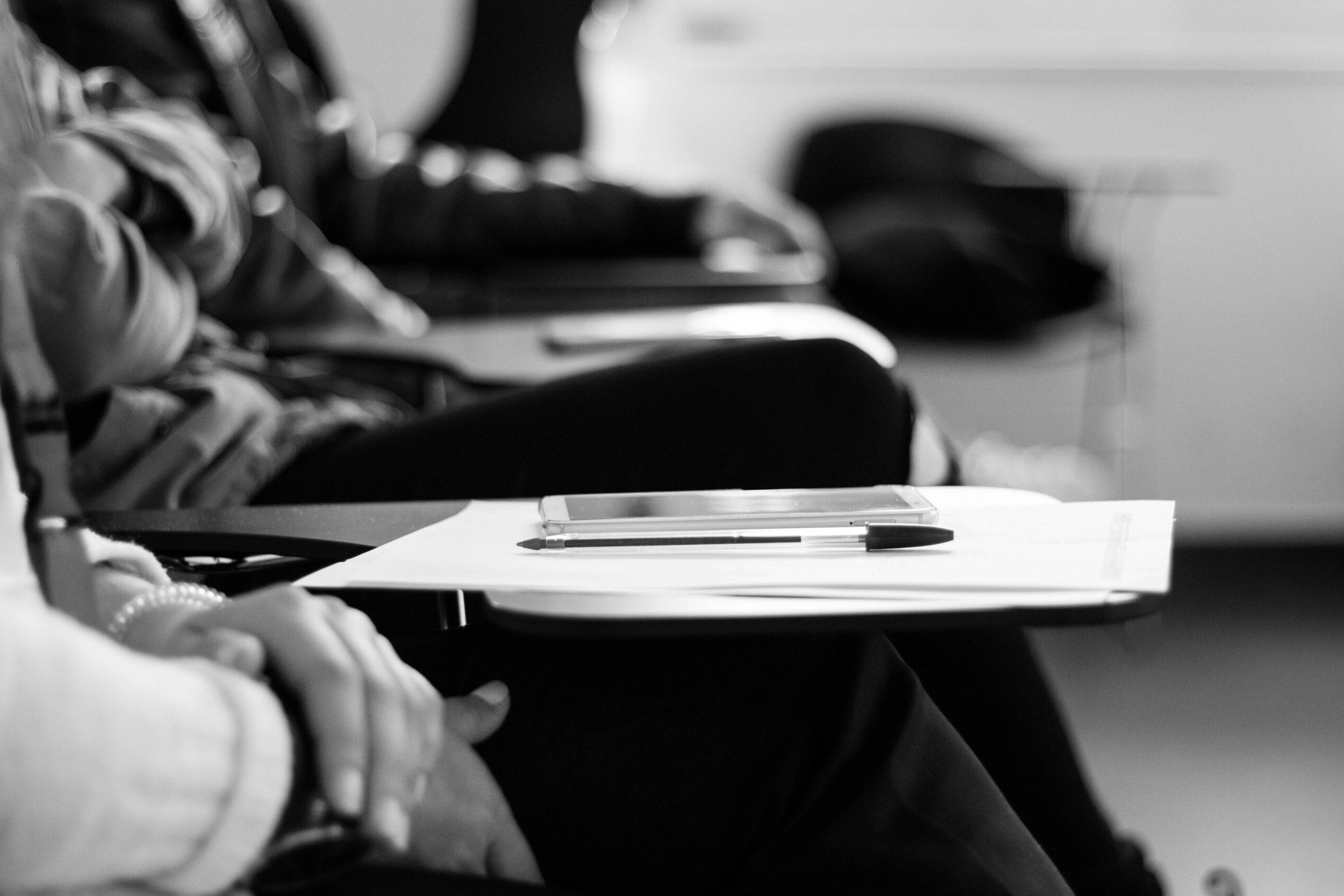 Black and white photograph of an individual at a school desk with paper, pen, and smart device on the desk